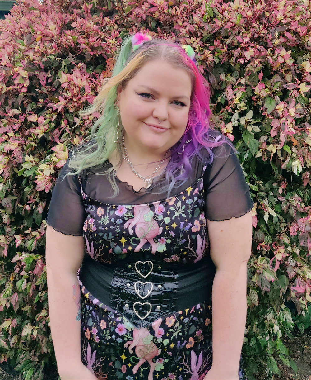 A chubby gal wearing a short sleeve lettuce hem mesh shirt with a midi length spaghetti strap dress in a floral and rabbit print dress. She wears silver mary jane shoes and a thick corset like beth with 3 silver hearts.
