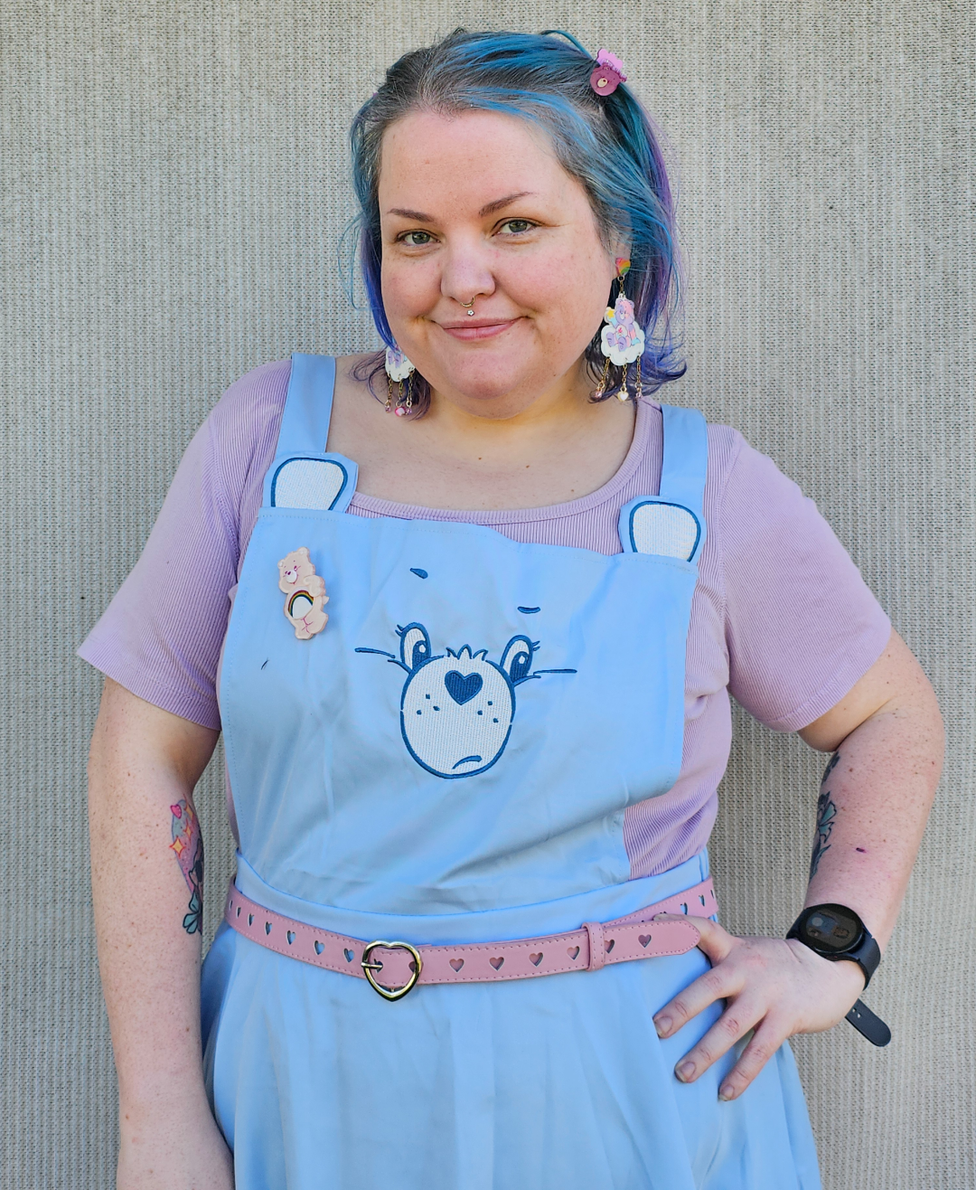 A picture of a chubby girl from the torso up. She has one arm down and the other resting on her tip. She has dusty blue shoulder length hair, clipped back with care bears hair clips. She is wearing a pastel purple short sleeved ribbed shirt, with a pastel blue care bears pinafore over. The pinafore has grumpy bears face on the front and ears at the straps. She wears a pink belt with love heart buckle and love heart holes.